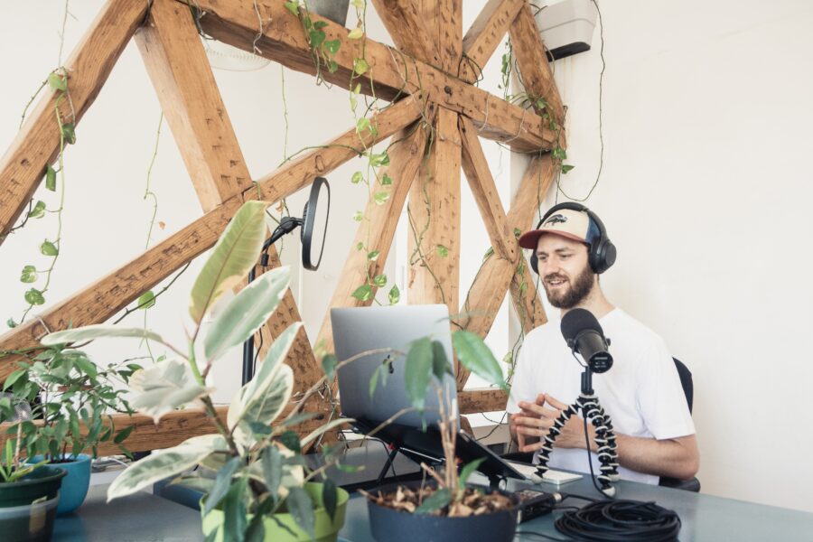 Male podcaster with laptop and equipment in front of him. There are plants on the desk, with wooden beams in the backdrop